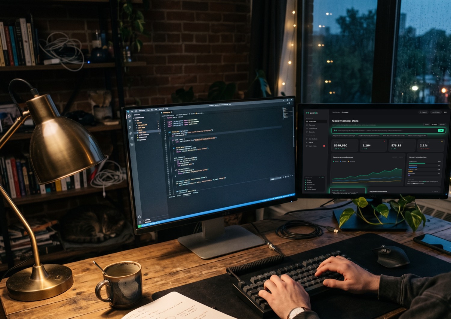 A late-night workspace: one monitor showing code, the other the live GoBrain dashboard, with hands at the keyboard, a notebook and coffee.